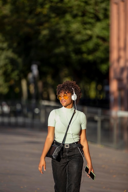 Morena jovem encaracolada de pele escura elegante em óculos de sol amarelos gosta de música em fones de ouvido