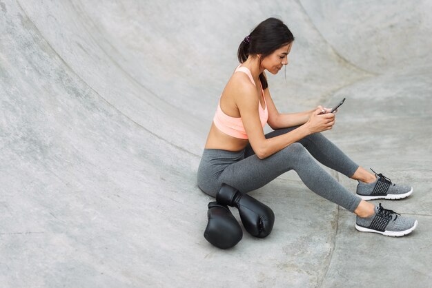 Morena joven en ropa deportiva sosteniendo el teléfono celular mientras está sentado en el piso de concreto con guantes de boxeo