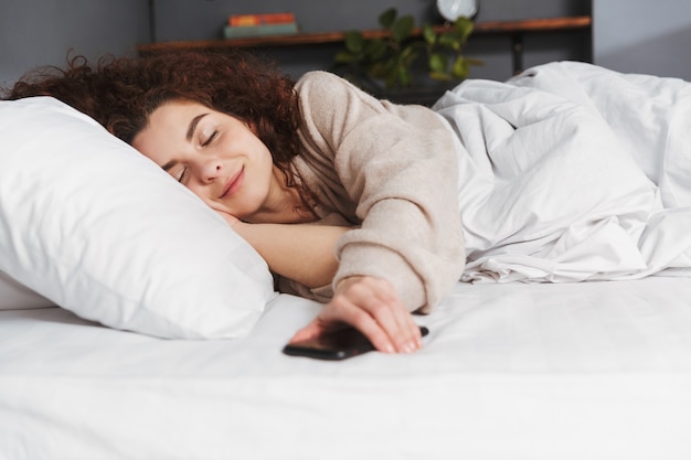 Morena joven vistiendo ropa de casa durmiendo con smartphone en la cama sobre ropa blanca en casa