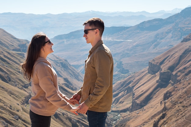 Morena con largo cabello suelto toma de la mano del hombre con gafas de sol de pie contra el profundo cañón entre altas montañas bajo la luz del sol