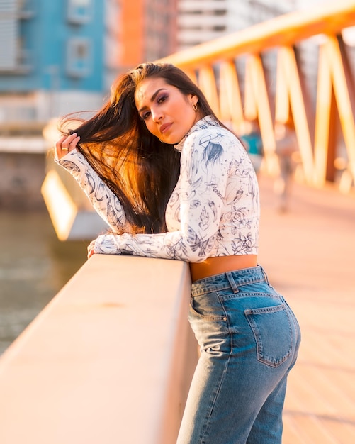 Una morena latina con jeans en la ciudad al atardecer En un puente mirando el río de la ciudad