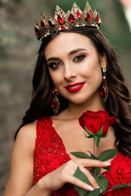 Morena linda em um vestido vermelho com uma coroa. menina morena com uma rosa vermelha do lado de fora na parede de pedra do loft