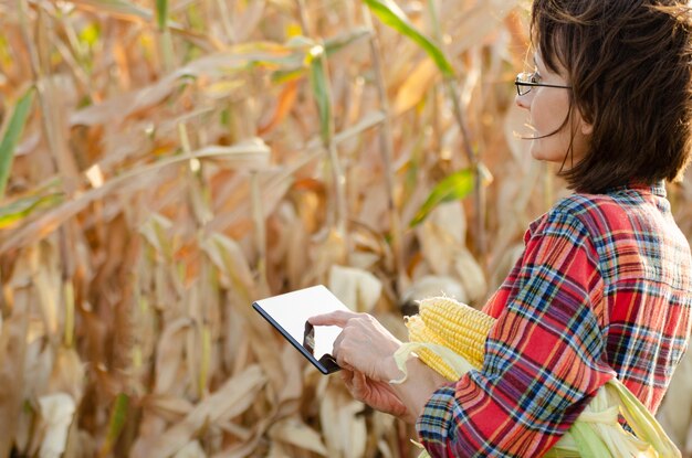 Morena de mediana edad trabajadora agrícola caucásica en gafas con tableta digital inspeccionando el campo de maíz día soleado de verano