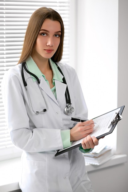 Morena médica sentada à mesa perto da janela no hospital e digitando no computador portátil