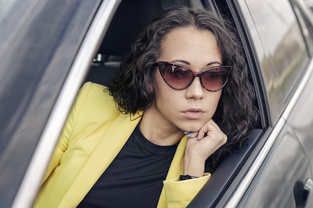 Morena motorista de óculos escuros, jaqueta amarela olha pela janela de seu carro.
