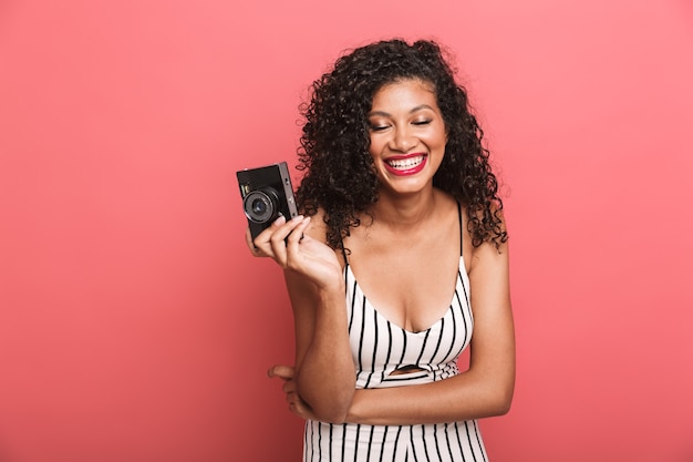 Morena mujer americana con cabello rizado fotografiando con cámara retro aislado sobre pared rosa