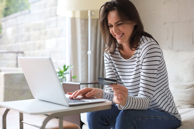 Foto morena natural feliz usando cartão de crédito e laptop na sala de estar.