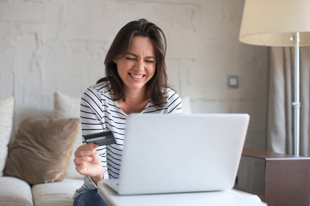 Morena natural feliz usando cartão de crédito e laptop na sala de estar.