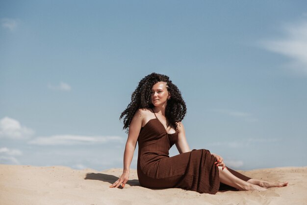 Morena oscura con cabello rizado voluminoso entre las arenas en una duna de arena con un vestido volador