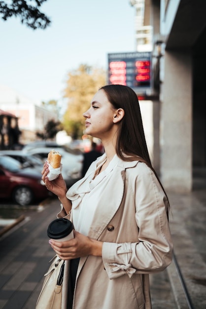 Morena de pelo largo con un abrigo ligero está comiendo pastel y café en una calle europea