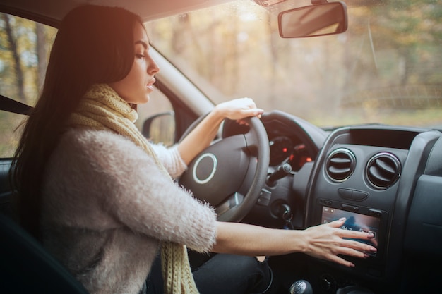 Morena de pelo largo en el auto. Una modelo femenina lleva un suéter y una bufanda. Concepto de otoño Viaje por el bosque otoñal en coche