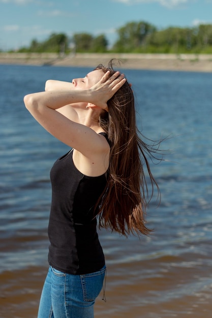 Morena de pelo largo en jeans de pie junto al mar y alisando su cabello en el viento