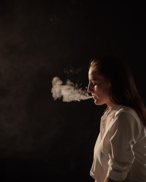 Foto morena pensativa con una camisa blanca fuma una cachimba en la oscuridad retrato de una mujer fumando un vape flotando fondo negro cigarrillo electrónico mal hábito