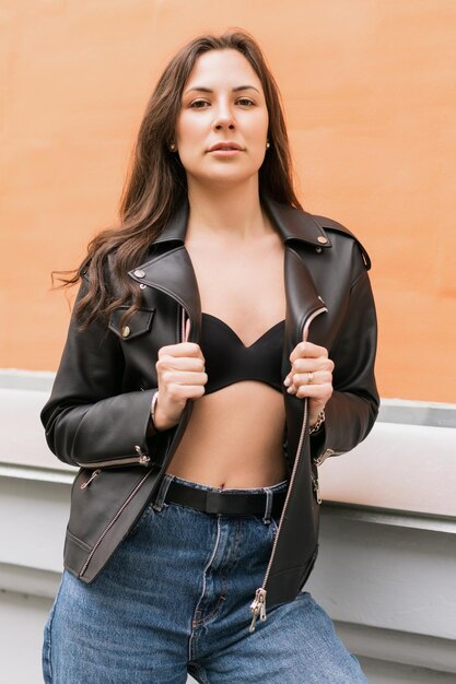Morena sexy con chaqueta de cuero y jeans Una mujer joven de buen humor posa en el fondo de un paisaje urbano