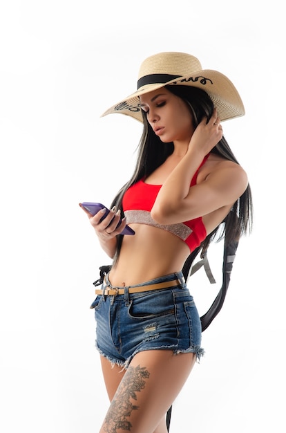 La morena con un sombrero en ropa de verano delgada y sexy con un teléfono aislado en un fondo blanco