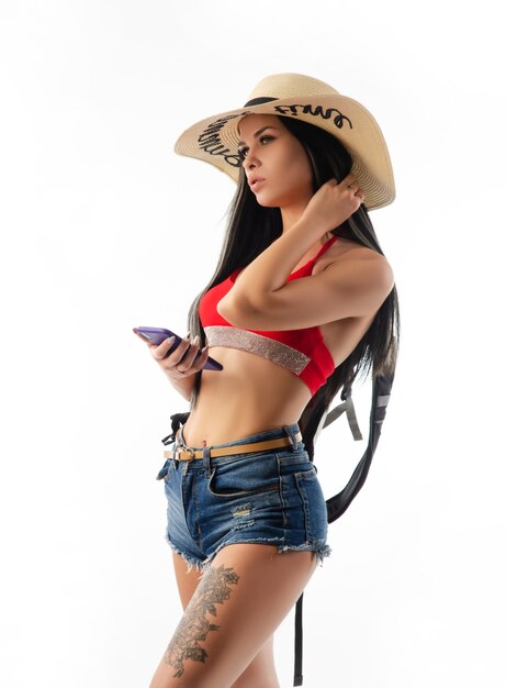 La morena con un sombrero en ropa de verano delgada y sexy con un teléfono aislado en un fondo blanco