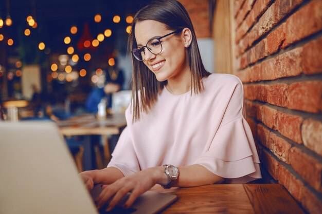 Morena sonriente con anteojos y elegante vestido sentado en un restaurante y usando una computadora portátil