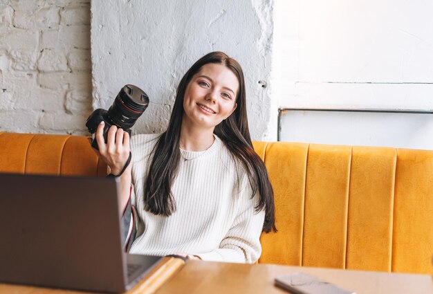 Morena sonriente joven fotógrafa trabajando con su cámara y portátil en el café