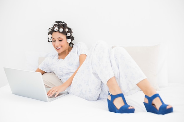 Morena sonriente en los rodillos de pelo y zapatos de cuña usando su computadora portátil en la cama