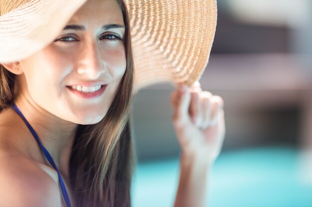 Foto morena sonriente sosteniendo sombrero junto a la piscina