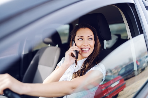 Morena sorridente caucasiana atraente, dirigindo o carro dela e falando ao telefone enquanto olha pela janela da calha.