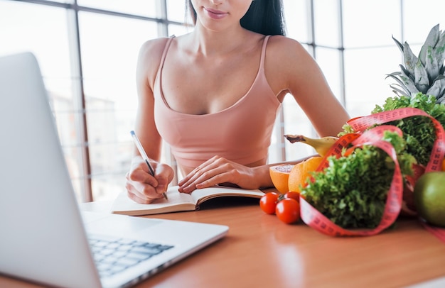 La morena con un tipo de cuerpo delgado se sienta junto a la mesa con una laptop y comida saludable.