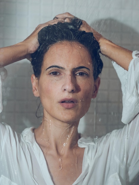 Foto morena vulnerável vestindo camisa branca coletando cabelo na cabine do chuveiro enquanto se molhava e olhava para a câmera