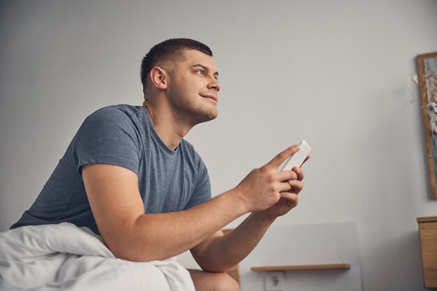 Moreno atraente com camiseta azul ficando na cama enquanto pensa e segura o celular nas mãos