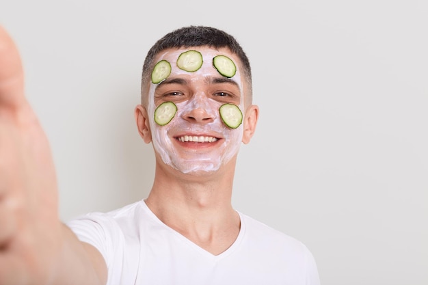 Un moreno feliz y satisfecho con una camiseta blanca haciendo una foto de punto de vista transmitiendo en vivo mientras realiza procedimientos cosméticos con máscara y pepinos aislados en un fondo gris