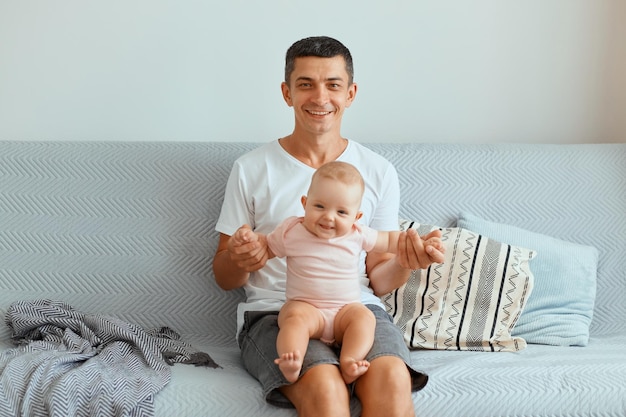 Moreno jovem bonito sentado no sofá em casa, segurando a filha infantil, brincando com uma garotinha encantadora, sorrindo, olhando para a câmera, vestindo roupas de estilo casual.