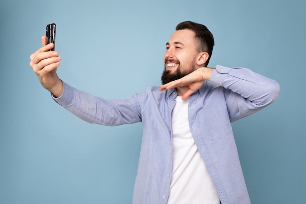 Moreno jovem e bonito barbudo vestindo roupas casuais elegantes, isolado na parede azul segurando um smartphone