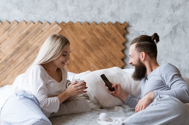 Morgen aufwachen Ritual. Lächelnder Mann und Frau im Bett mit Getränk und Smartphone.