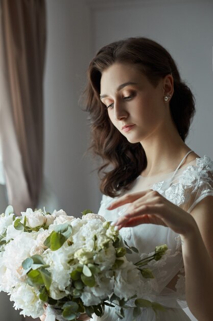 Morgen der Braut. Eine schöne Frau bereitet sich auf eine Hochzeit, natürliches Make-up und eine schicke Frisur vor. Weißes Peignoir und das Hochzeitskleid