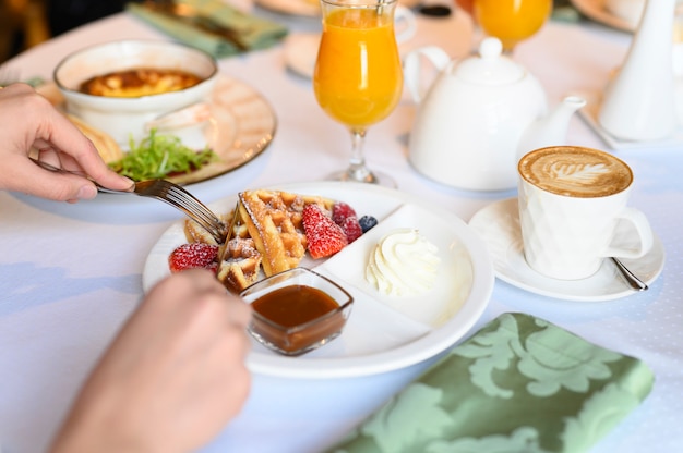 Morgen Frühstück oder Brunch im Restaurant. Tisch mit Getränken und Essen. Frauenhände schneiden Wiener Waffeln mit Messer und Gabel. selektiver Fokus