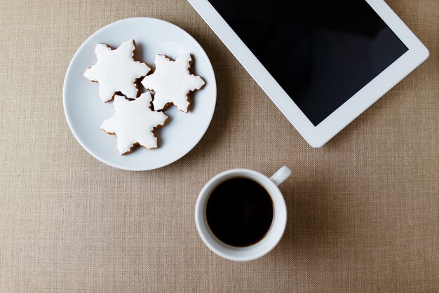 Morgen Tasse Kaffee auf dem schäbigen weißen Schreibtisch mit digitaler Tablette mit leerem Bildschirm, Kerze, frischem Keks