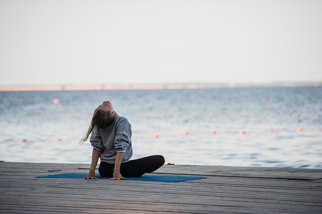 Morgenaufnahme eines Mädchens in Lotussitz, das Stretchyogaübungen auf dem Pier macht