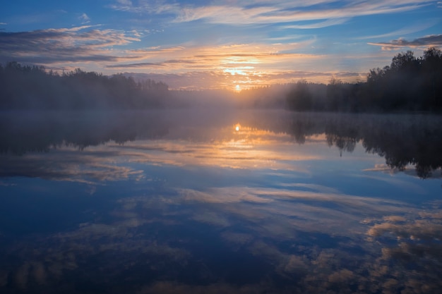 Morgendämmerung über einem nebligen See im Wald am frühen Morgen. Blauer Himmel und Sonne spiegelten sich im Wasser