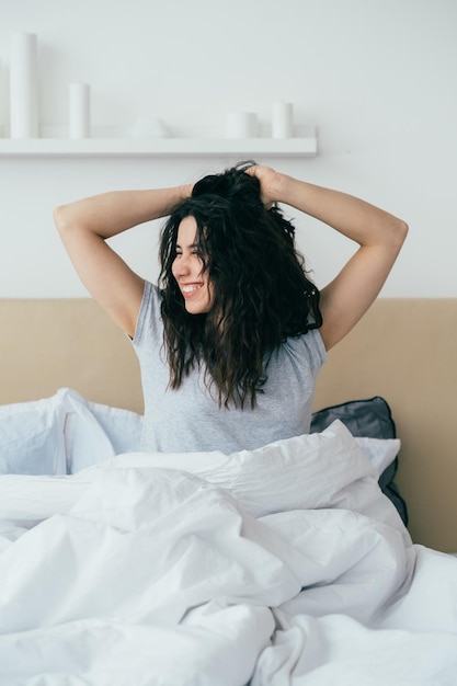 Morgenenergie Heimwochenende Neuanfangsmotivation Fröhlich inspirierte brünette Frau, die sich lächelnd im Bett im Schlafzimmer mit Sonnenlicht ausdehnt