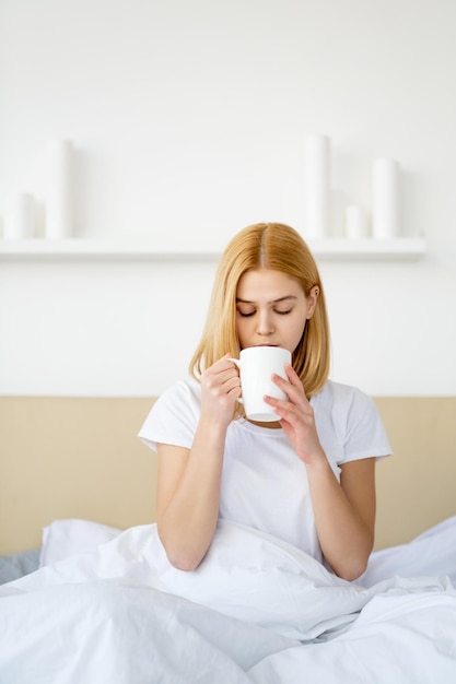 Morgenkaffee Schlafenszeit Freizeit Fauler Sonntag Wochenendruhe Friedliche junge Frau unter Decke in weichen weißen Bettlaken, die eine Tasse Tee im Bett im hellen, gemütlichen Schlafzimmer genießt