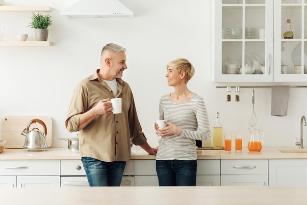 Morgenkaffee, Wohnkomfort, Familienfrühstück und glückliche Beziehungen. Erwachsener ziemlich positiv lächelnder Ehemann und Ehefrau, die heißen Tee in einem gemütlichen modernen Kücheninterieur sprechen und trinken, leerer Raum