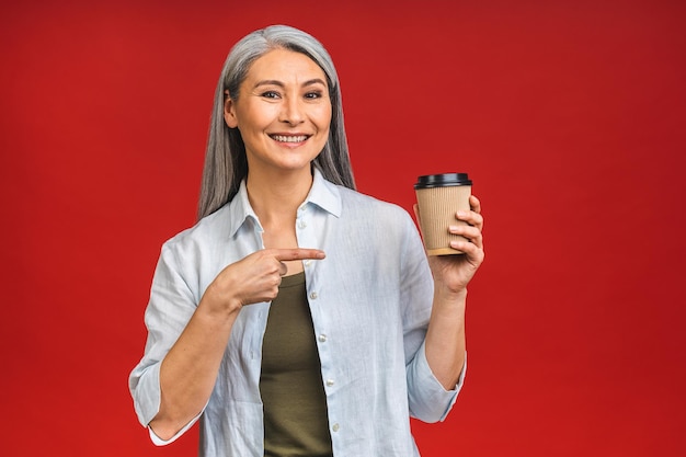 Morgenkaffee zum Mitnehmen Charmante Seniorin in formeller Kleidung, die mit einem Pappbecher mit heißem Getränk steht, das auf rotem Hintergrund isoliert ist und sich auf einen guten Arbeitstag vorbereitet Reife Geschäftsfrau macht eine Kaffeepause