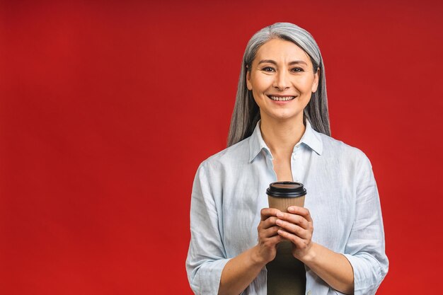 Morgenkaffee zum Mitnehmen Charmante Seniorin in formeller Kleidung, die mit einem Pappbecher mit heißem Getränk steht, das auf rotem Hintergrund isoliert ist und sich auf einen guten Arbeitstag vorbereitet Reife Geschäftsfrau nimmt eine KaffeepausexA