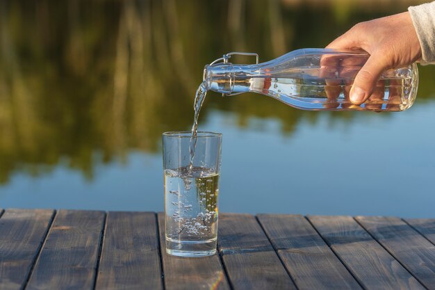 Morgens neben dem See und dem Wald im Frühjahr aus einer Flasche Wasser in ein Glas gießen, Nahaufnahme. Natur- und Reisekonzept