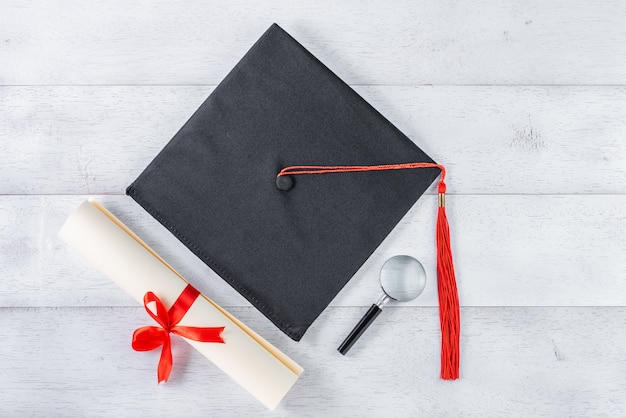 Mortarboard, lupa e diploma amarrado com fita vermelha na mesa de madeira branca, vista superior