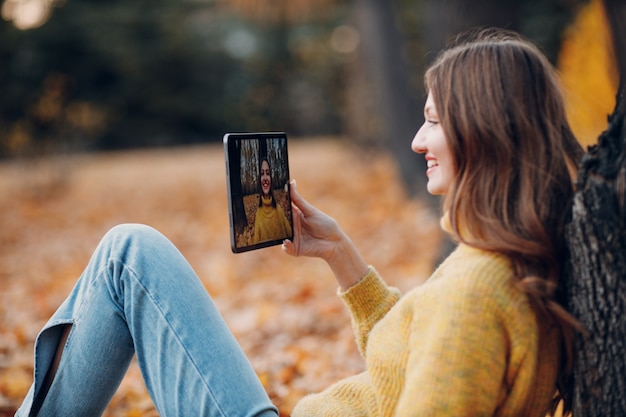 Moscú, Rusia - 19 de octubre de 2021: modelo de mujer joven tomar foto selfie con tablet pc en el parque de otoño con hojas de arce de follaje amarillo. Moda de temporada de otoño