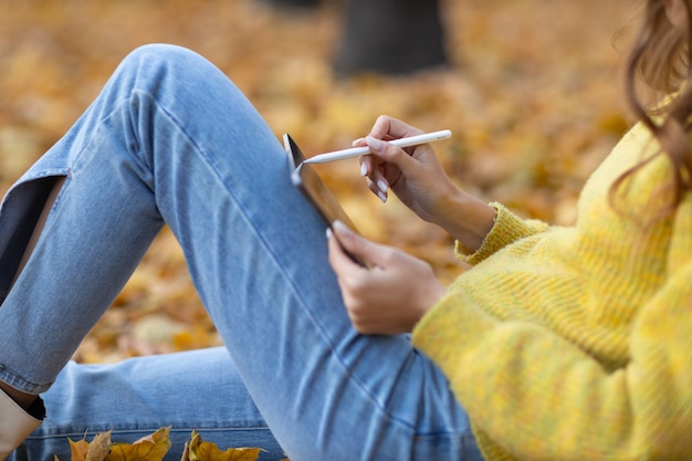 MOSKAU, RUSSLAND - 19. OKTOBER 2021: Junges Frauenmodell, das mit Tablet-PC sitzt und mit Bleistiftstift im Herbstpark mit gelben Ahornblättern malt.