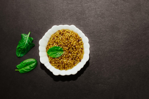 Mostaza de grano con una hoja verde en un recipiente blanco sobre un fondo negro