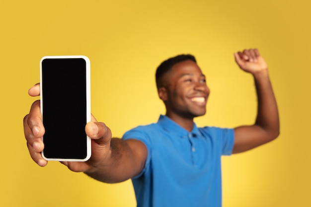 Mostra a tela do telefone, vencedor. Retrato de jovem afro-americano isolado no fundo amarelo do estúdio, expressão facial. Lindo modelo masculino, copyspace. Conceito de emoções humanas, vendas, anúncio.