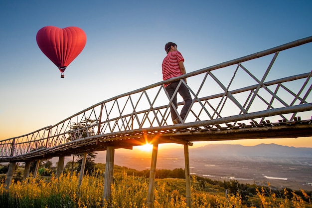 Mostrado em silhueta do homem asiático que está na ponte de bambu com o balão de ar quente vermelho na forma de um coração sobre o por do sol.