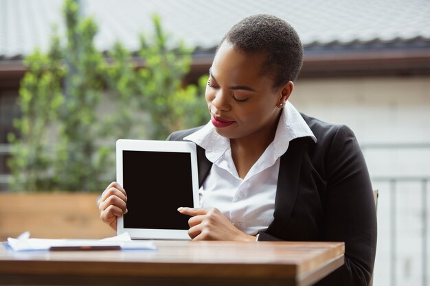 Mostrando a tela do tablet em branco. Mulher de negócios afro-americana em traje de escritório, sorrindo, parece confiante, ocupada. Finanças, negócios, igualdade, conceito de direitos humanos. Bela jovem modelo feme, bem-sucedida.